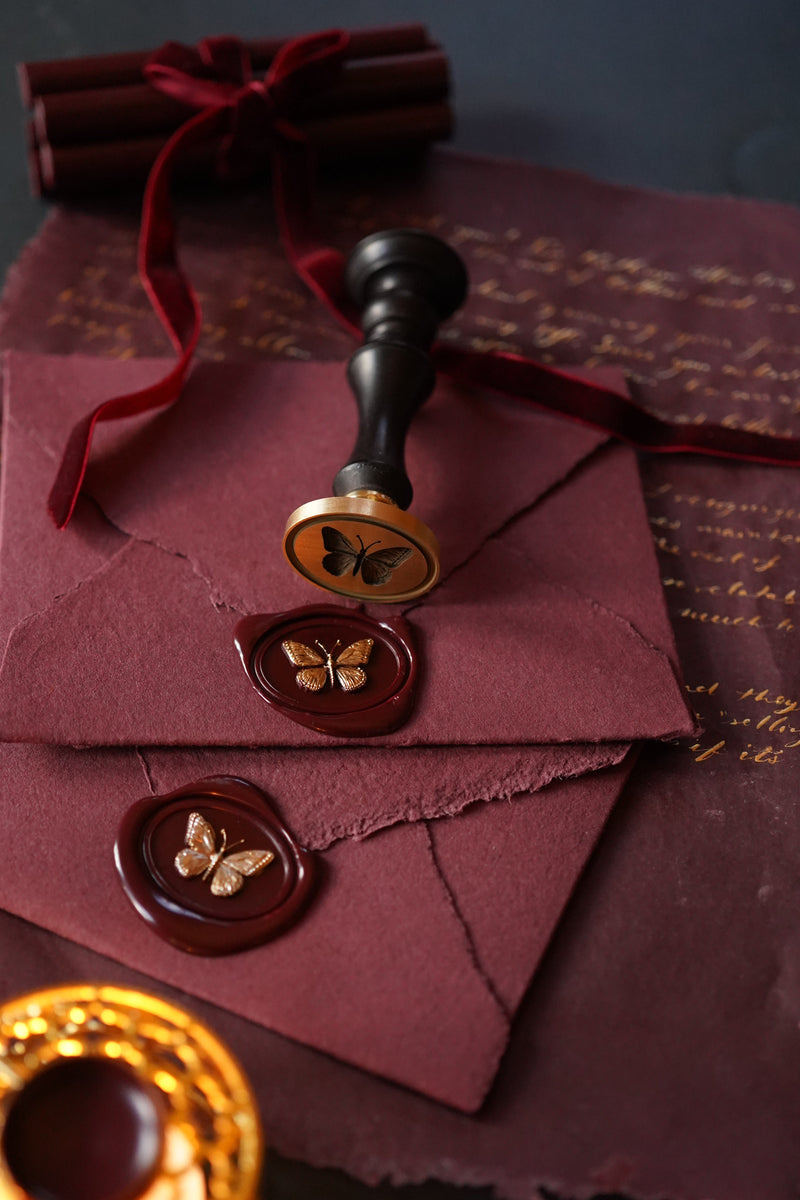 A styled flat-lay of luxury wax seals of butterflies hand-painted gold and envelopes. Two dark burgundy paper envelopes are sealed with round dark red wax stamps featuring a gold butterfly motif, one with a black-handled wax seal and one with a gold-handled wax seal.